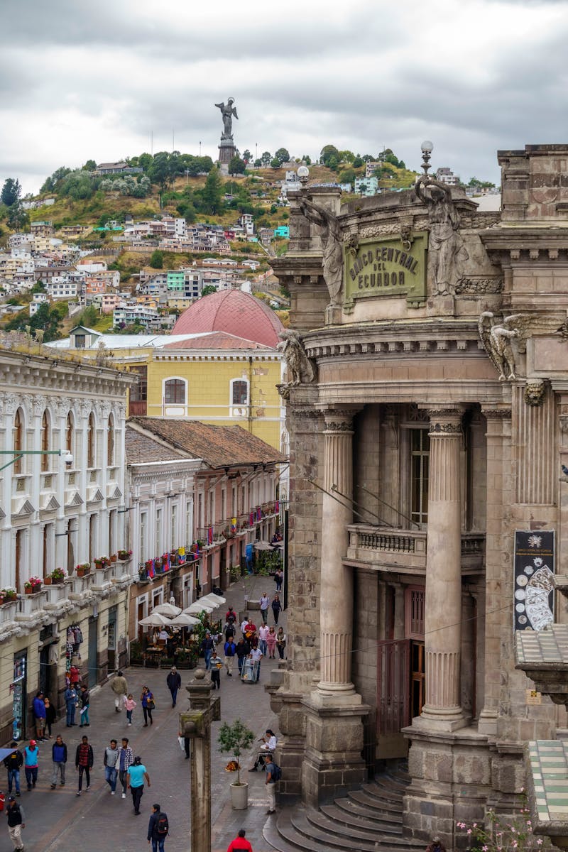 Capture of a bustling street in Quito with a view of El Panecillo statue on the hill, showcasing rich architecture.